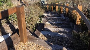 Un escalier en bois mène à une plage d’où l’on a une vue dégagée sur l’océan, avec une rangée de bâtiments blancs en arrière-plan.