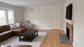 A living room with a brown leather sofa, a dark coffee table, a white armchair, a fireplace, and a TV mounted on the wall. - Skyline View Home w/ Office - Minutes From Culver, LAX, Santa Monica & SOFI (Los Angeles)