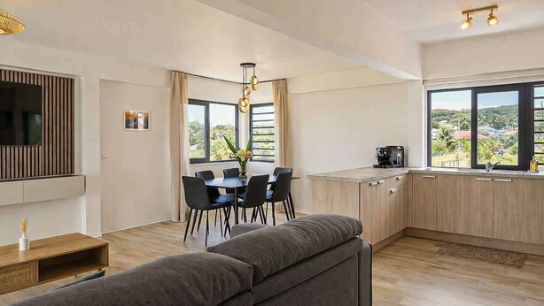 A modern living room with a sofa, dining table, and chairs. There is a kitchen area with cabinets and a coffee machine. Large windows provide a view of the outdoors. - New 2BR Apartment with AC Near Lagoon & Dining (Taiarapu)
