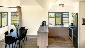 A modern kitchen with a central island, black chairs, a dining table, and large windows offering a view of the outdoors. - New 2BR Apartment with AC Near Lagoon & Dining (Taiarapu)