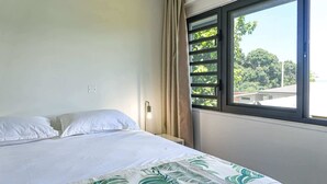 A single bed with white linens, a window with a view of greenery and buildings, and a curtain on the window. - New 2BR Apartment with AC Near Lagoon & Dining (Taiarapu)