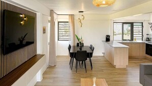 A modern living room with a dining area, a kitchen, and a large TV mounted on the wall. - New 2BR Apartment with AC Near Lagoon & Dining (Taiarapu)