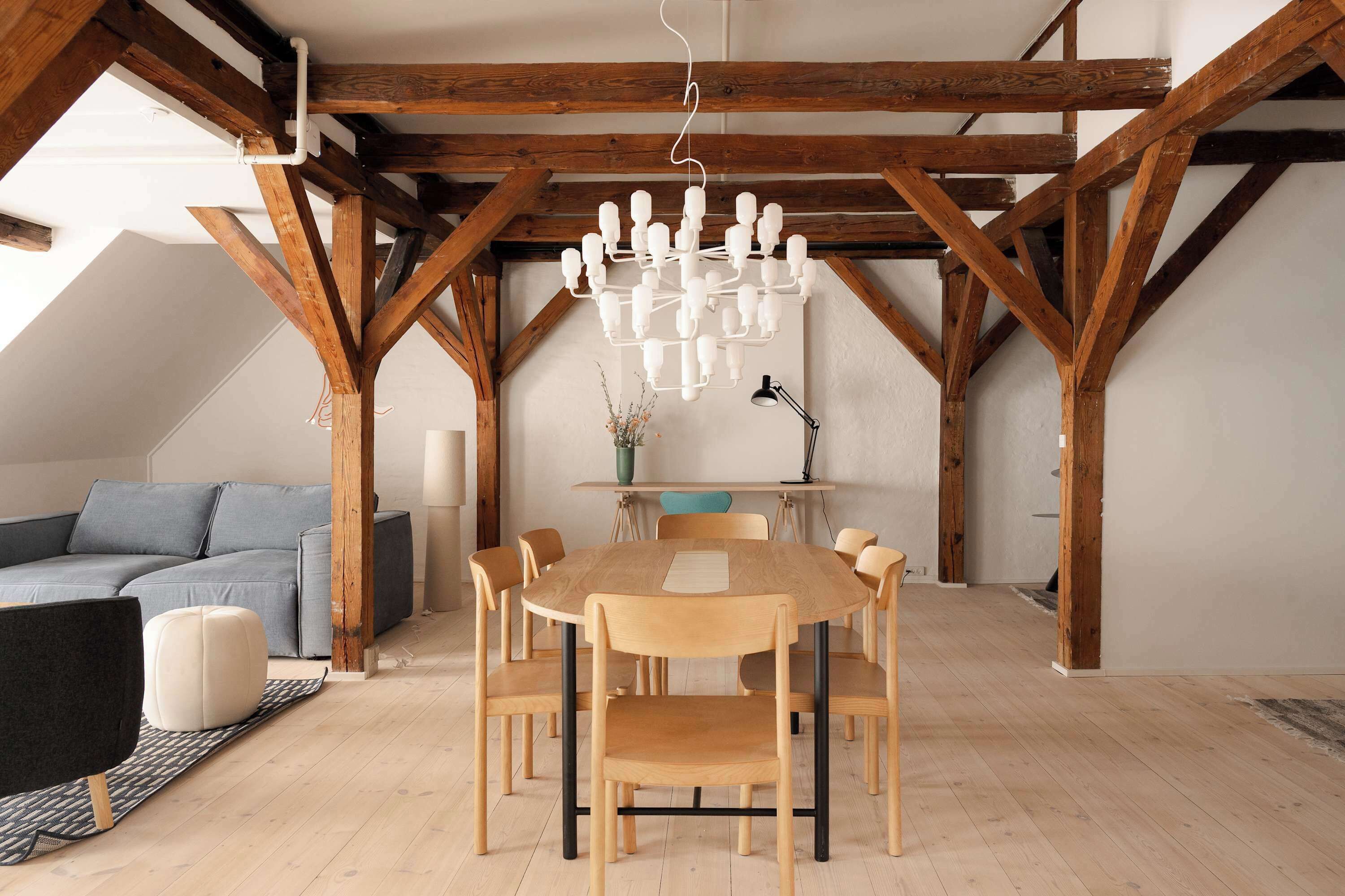 A dining area with a wooden table and chairs, a chandelier, and a blue sofa in a loft-style room with exposed wooden beams.
