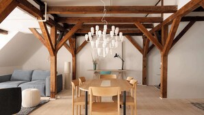 Een eetgedeelte met een houten tafel en stoelen, een kroonluchter en een blauwe bank in een loft-achtige kamer met zichtbare houten balken.