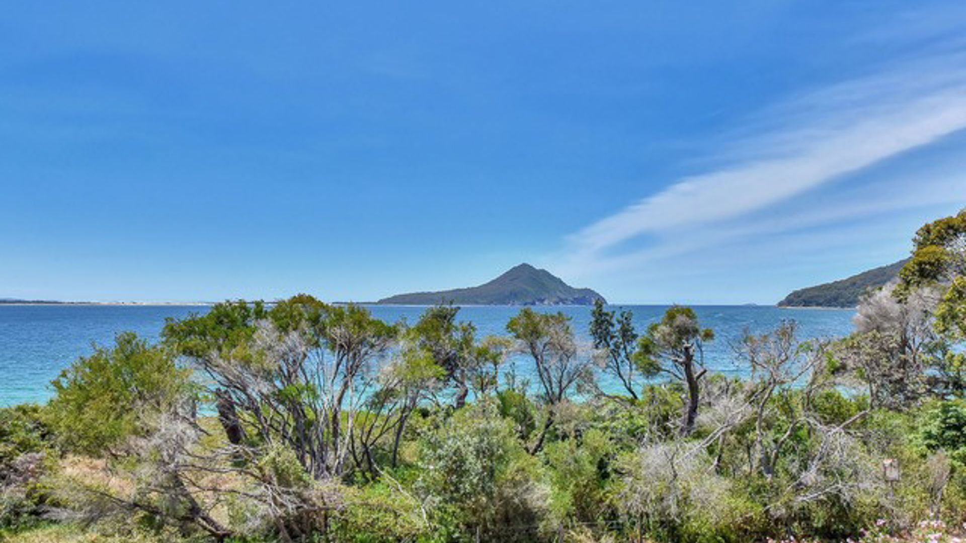 A coastal landscape with clear blue waters, a prominent mountain in the distance, and dense greenery in the foreground.