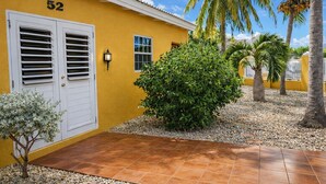 Un edificio amarillo con el número '52' en la pared, una puerta blanca con contraventanas, un pequeño arbusto y una palmera.