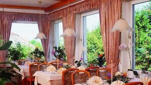 Un'area pranzo con tavoli apparecchiati, sedie e ampie finestre con tende.