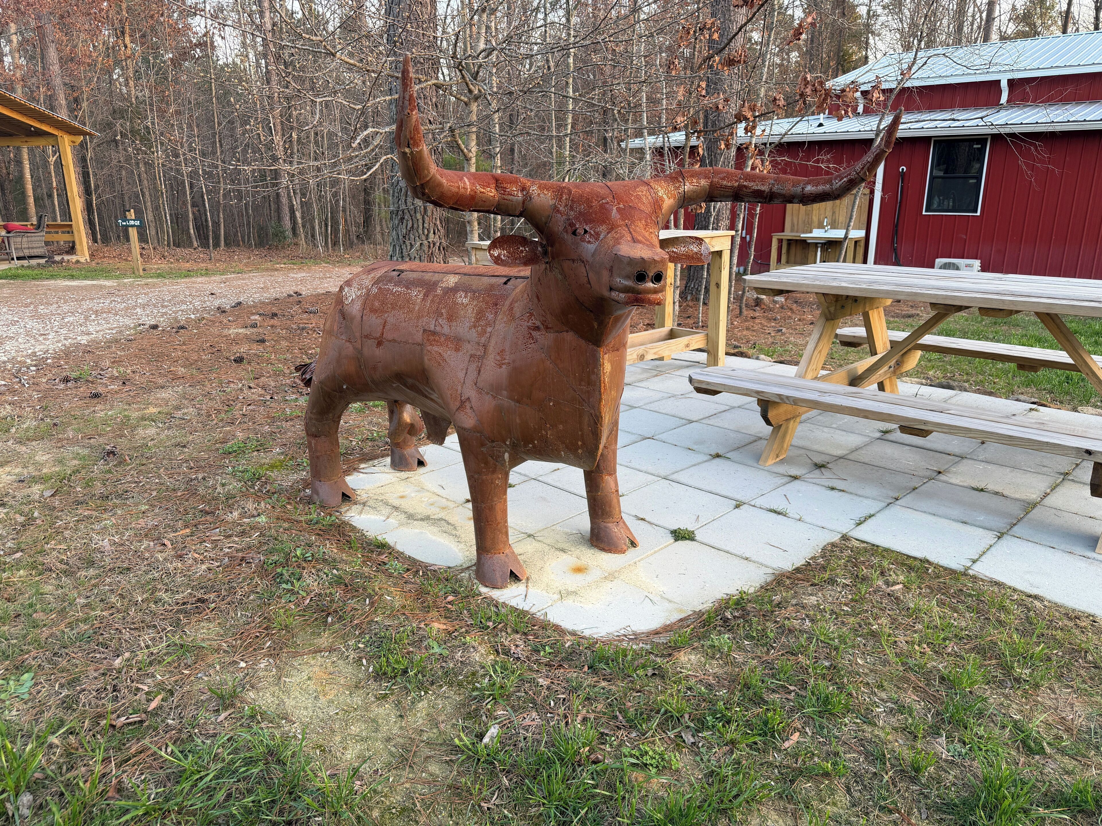 A rustic outdoor area with a large metal bull sculpture, a picnic table, and a red building in the background.