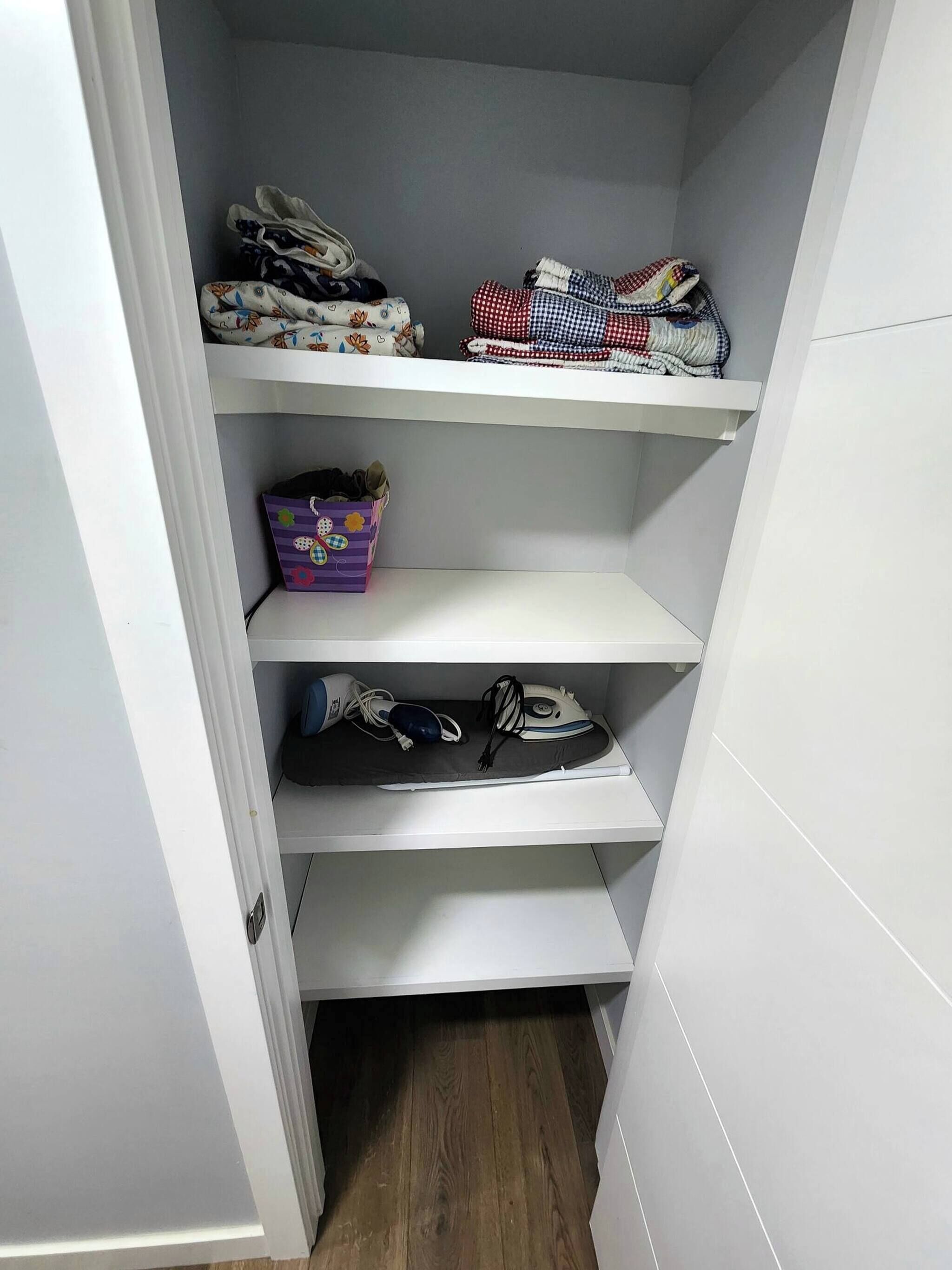Un placard avec des étagères où sont pliés des vêtements, une boîte de rangement violette et une planche à repasser avec un fer à repasser et un sac bleu.