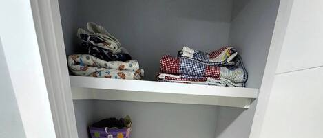 Un placard avec des étagères où sont pliés des vêtements, une boîte de rangement violette et une planche à repasser avec un fer à repasser et un sac bleu.