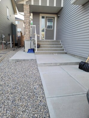 A residential building with a stone pillar, a gray door, and a set of concrete steps leading to a porch. - Luxurious Home Away From Home With Aircon (Airdrie)