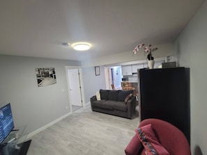 A living room with a grey sofa, a red chair, a flat-screen TV, and a kitchen area in the background.