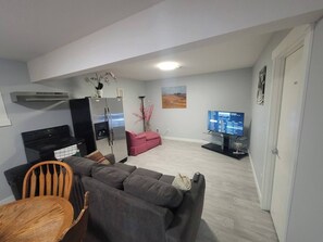 A modern living room with a grey sofa, a wooden chair, a black TV stand, a pink sofa, and a refrigerator. - Luxurious Home Away From Home With Aircon (Airdrie)
