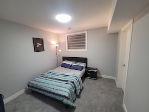 A bedroom with a bed, a bedside table, a lamp, a framed picture, and a window with blinds.