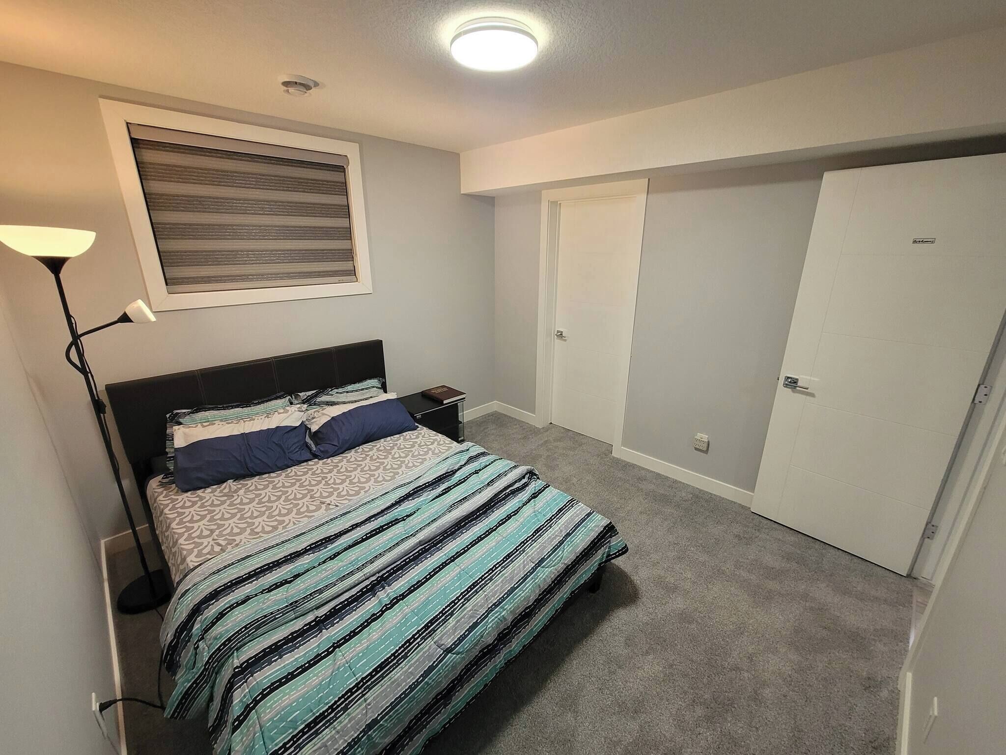 A bedroom with a bed, a nightstand, a lamp, a window with blinds, and a closet.