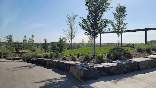 A landscaped area with a paved walkway, a grassy hill, and a metal structure in the background. - Luxurious Home Away From Home With Aircon (Airdrie)