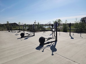An outdoor exercise area with various fitness equipment, including weight machines and benches, set against a backdrop of trees and a distant view of a lake. - Luxurious Home Away From Home With Aircon (Airdrie)