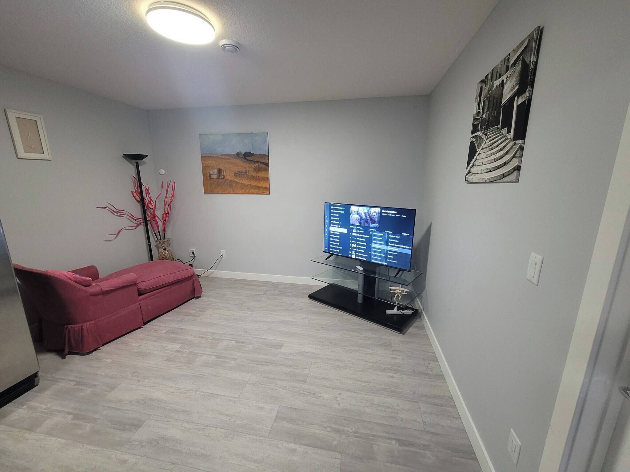 A living room with a flat-screen TV on a stand, a red sofa, a painting on the wall, and a floor lamp.