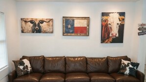 A brown leather sofa with a wooden coffee table in front, three artworks on the wall, and a window with blinds. - Saddle Up at Rodeo Ranch House! Cowboy Vibes, Blocks from the Stockyards Action! (Fort Worth)