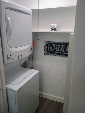 A compact laundry room with a front-loading washer and dryer, a small shelf with a container, and a decorative sign on the wall. - Saddle Up at Rodeo Ranch House! Cowboy Vibes, Blocks from the Stockyards Action! (Fort Worth)