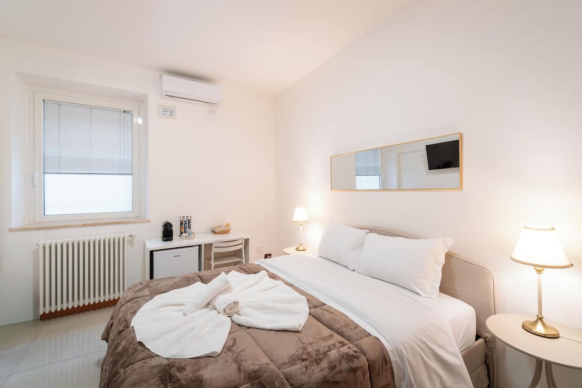 Una habitación de hotel con cama, lámparas de noche, un escritorio con silla, un radiador y una ventana con persianas.