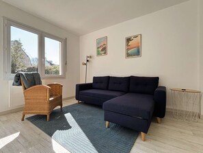 Un salon moderne avec un canapé d’angle bleu foncé, un fauteuil en osier, une petite table d’appoint ronde et deux tableaux encadrés accrochés au mur.
