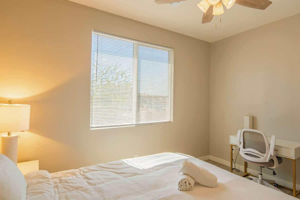 A hotel room with a bed, a desk, a chair, a window with blinds, and a ceiling fan.