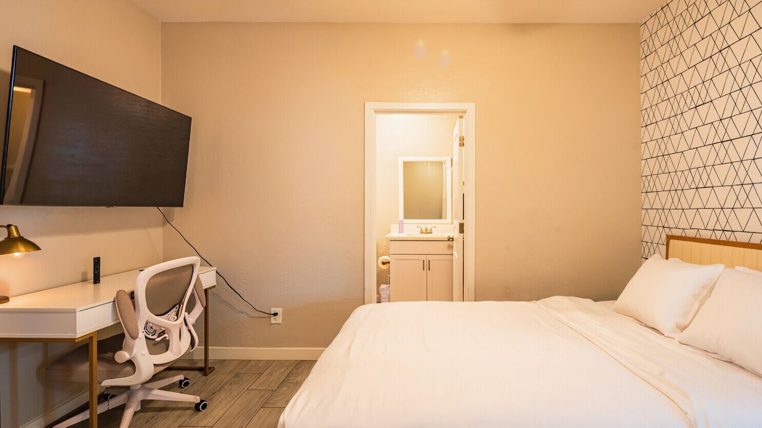 A hotel room with a bed, a desk with a chair, a TV mounted on the wall, and a door with a mirror.