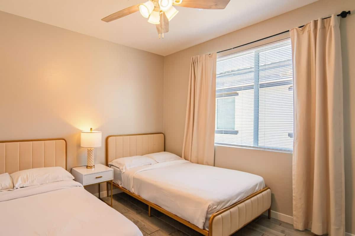 A bedroom with two beds, a ceiling fan, a bedside table with a lamp, and a window with curtains.