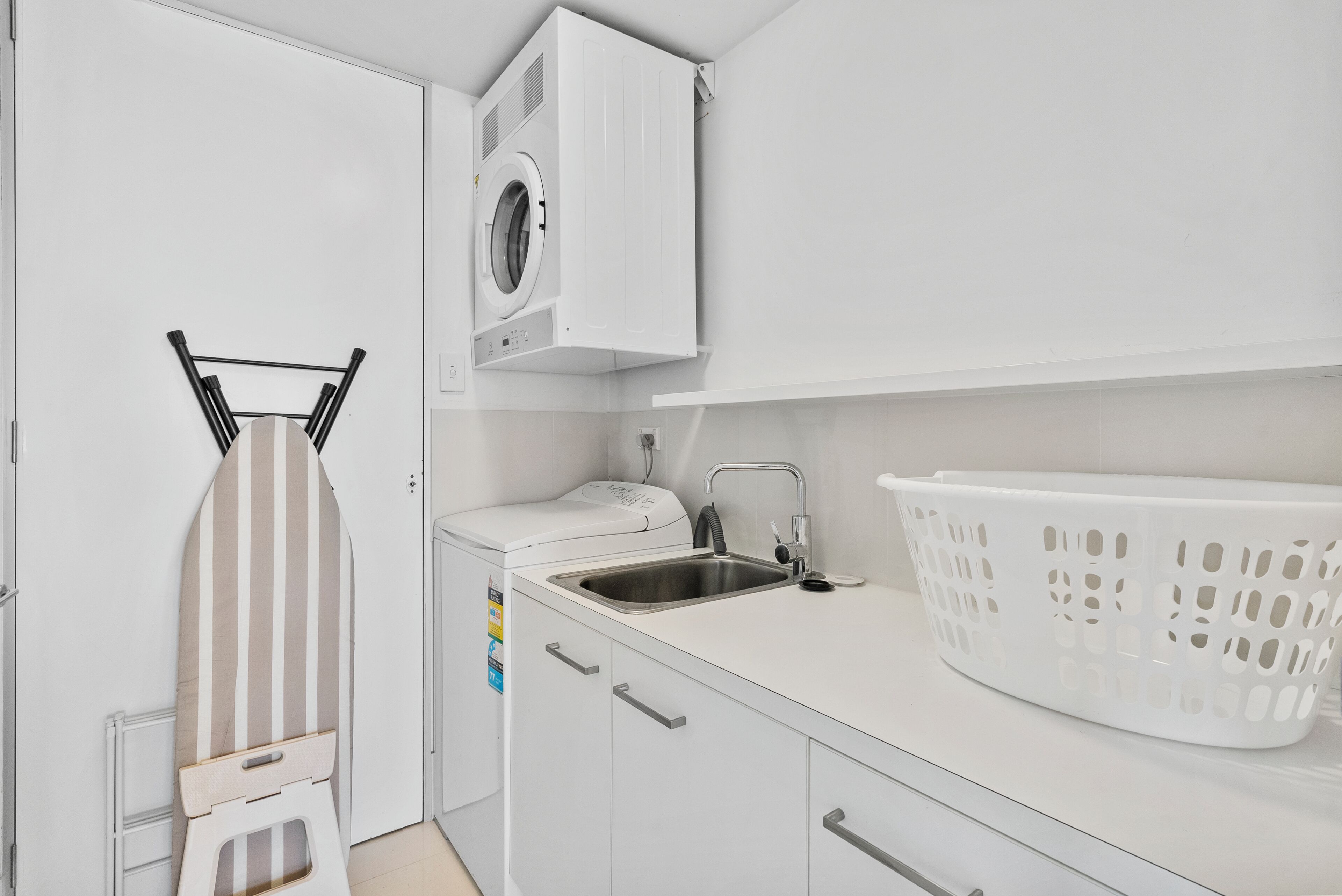 A compact laundry area with a washing machine, a drying rack, a sink, and a laundry basket.