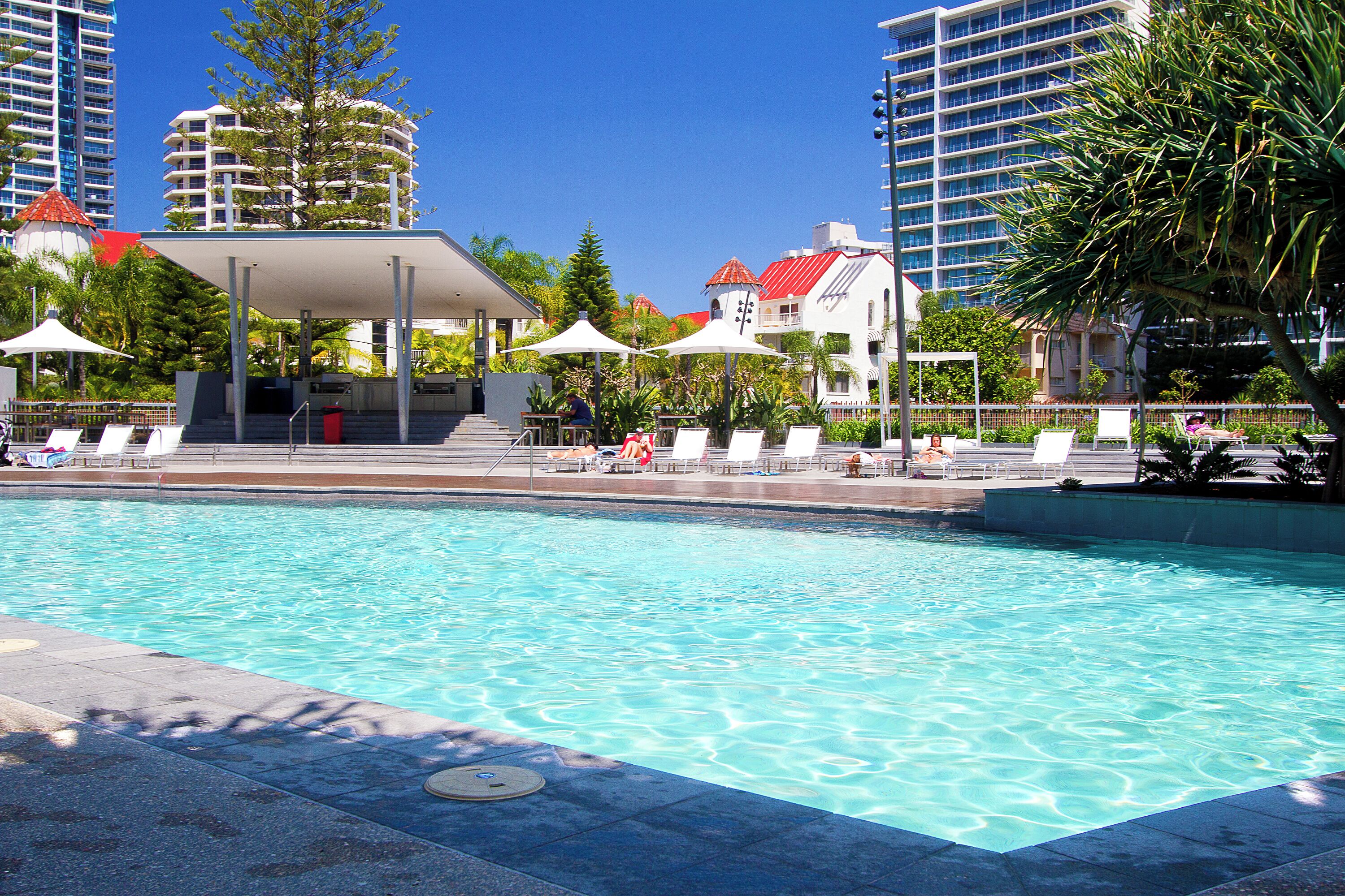 Una piscina con sillas de descanso y sombrillas, rodeada de edificios altos y árboles.