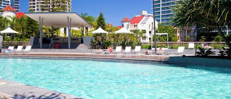 Una piscina con sillas de descanso y sombrillas, rodeada de edificios altos y árboles.