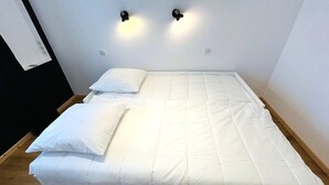 Uma cama de solteiro com roupa de cama branca em um quarto com parede branca lisa e duas luminárias fixadas na parede.