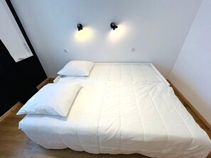 En enkeltseng med hvide sengetøj i et rum med en ensfarvet, hvid væg og to vægmonterede lamper.