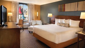 A hotel room with a large bed, a desk with a TV, two chairs, a window with curtains, and a city view.