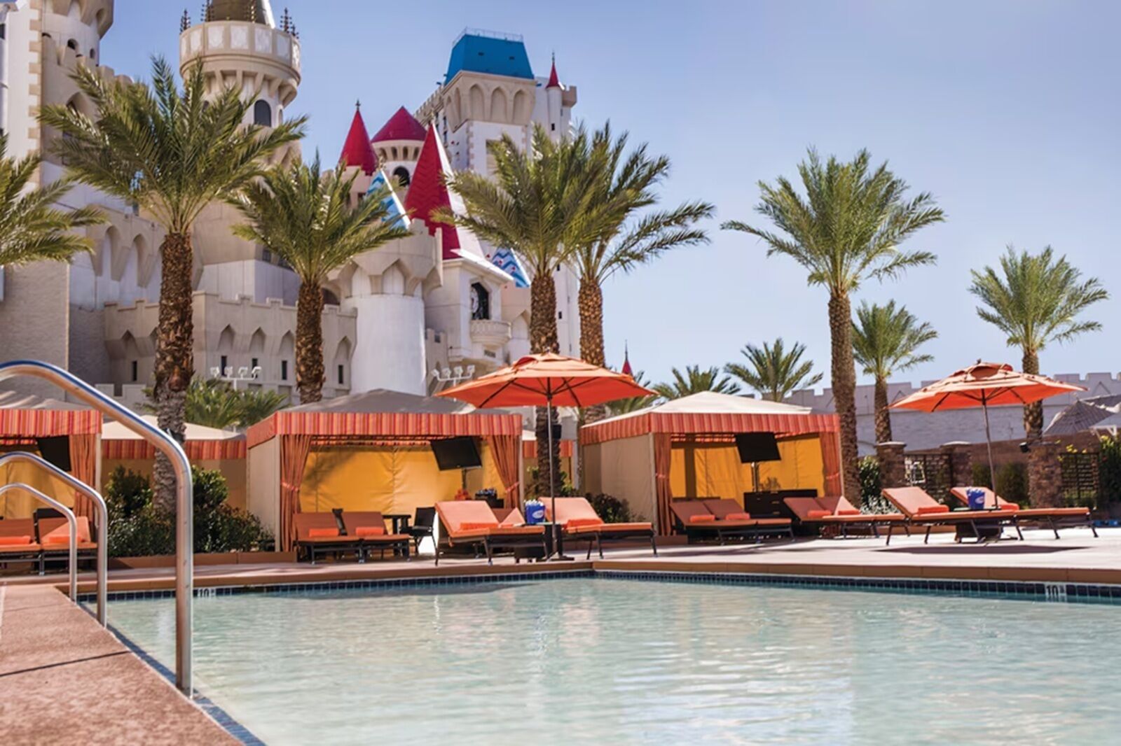 A pool area with lounge chairs and umbrellas, palm trees, and a castle-like building in the background.