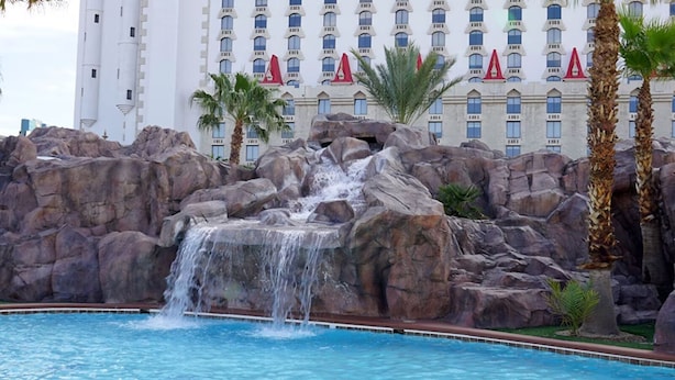 A pool with a waterfall feature surrounded by large rocks and palm trees, with a hotel building in the background.