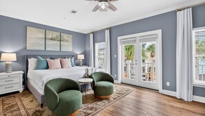 A bedroom with a bed, two green armchairs, a small round table, a ceiling fan, and a view of the outdoors through a sliding glass door. - Sun Sand Repeat - Modern Coastal Townhome in Gulf Place (Santa Rosa Beach)