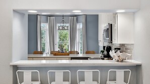 A modern kitchen with a central island, white bar stools, and a view of greenery through the window. - Sun Sand Repeat - Modern Coastal Townhome in Gulf Place (Santa Rosa Beach)