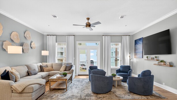 A modern living room with a beige sectional sofa, a coffee table, and blue armchairs. There is a ceiling fan, a flat-screen TV, and a large window with curtains. - Sun Sand Repeat - Modern Coastal Townhome in Gulf Place (Santa Rosa Beach)