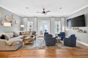A modern living room with a beige sectional sofa, a coffee table, and blue armchairs. There is a ceiling fan, a flat-screen TV, and a large window with curtains. - Sun Sand Repeat - Modern Coastal Townhome in Gulf Place (Santa Rosa Beach)