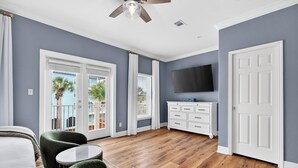 A room with a wooden floor, a ceiling fan, a flat-screen TV, a white dresser, and a balcony with a view of palm trees. - Sun Sand Repeat - Modern Coastal Townhome in Gulf Place (Santa Rosa Beach)