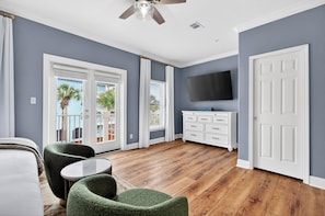 A room with a wooden floor, a ceiling fan, a flat-screen TV, a white dresser, and a balcony with a view of palm trees. - Sun Sand Repeat - Modern Coastal Townhome in Gulf Place (Santa Rosa Beach)