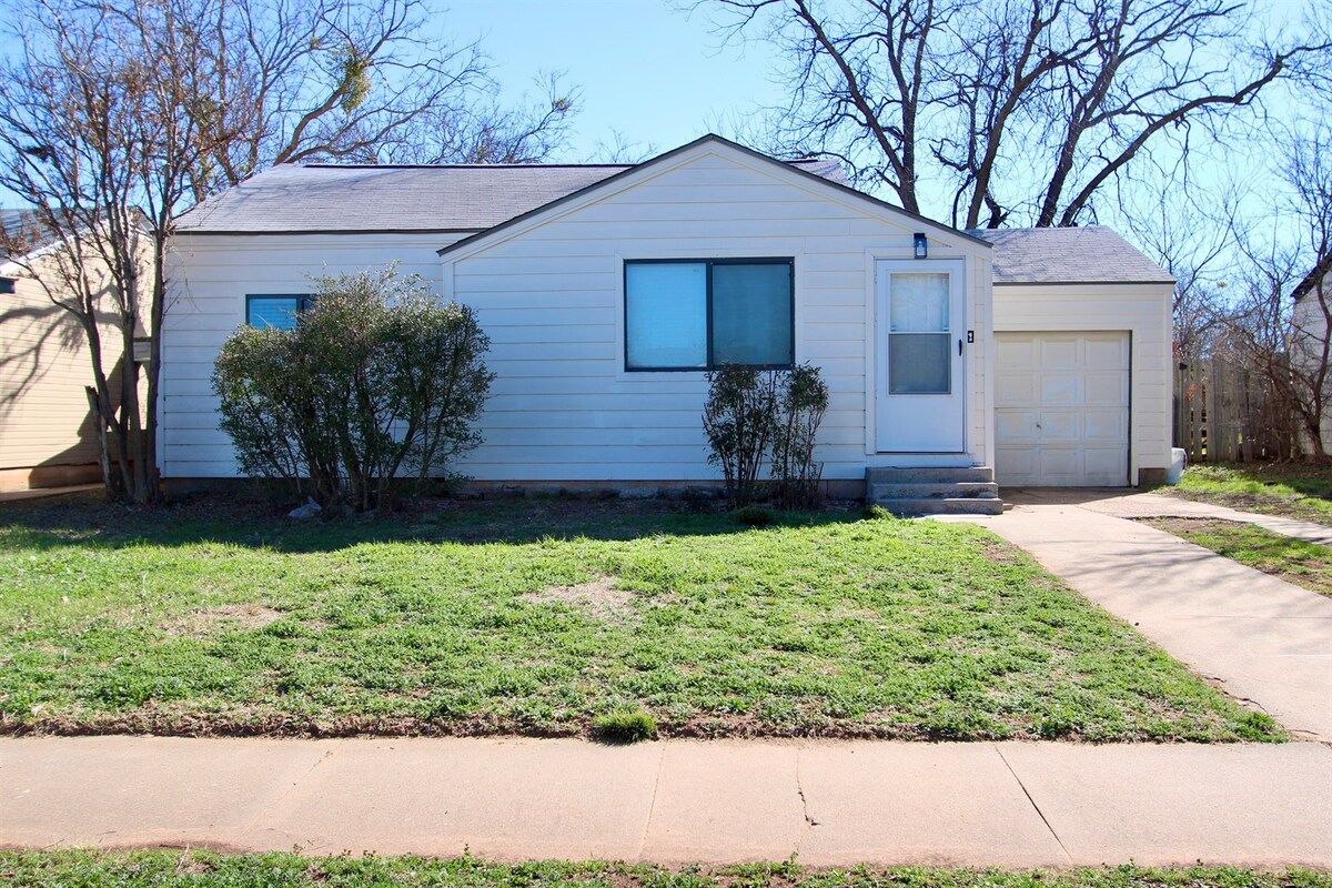 A single-story house with a garage, a front yard with grass and a small tree, and a clear sky.
