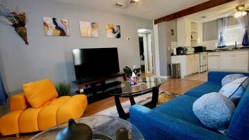 A living room with a sofa, a coffee table, and a television. There is a kitchen area visible in the background.