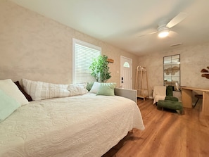 A bedroom with a large bed, a desk, a chair, a plant, and a mirror on the wall.