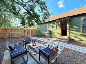 A patio with outdoor furniture and a small table, surrounded by a wooden fence and a green house.