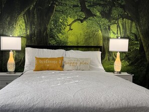 Uma cama com papel de parede com tema de floresta, duas luminárias de cabeceira e duas almofadas com texto.