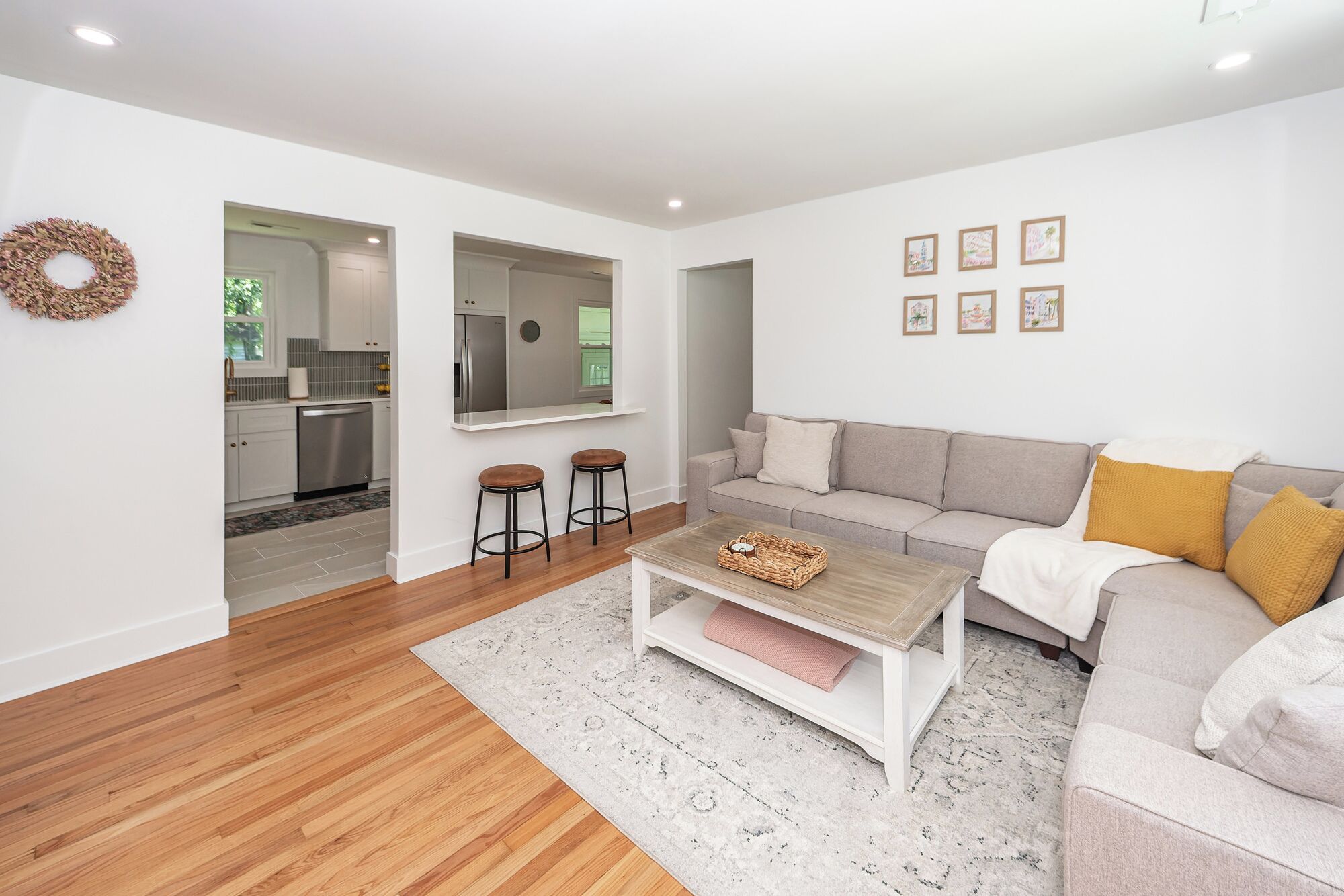 A modern living room with a sofa, coffee table, and two bar stools. There is a kitchen visible through an open doorway.