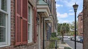 A brick building with red shutters, a small porch, and a street view with parked cars and palm trees. - Savannah Home, Sleeps 6, Prime Location (Savannah)